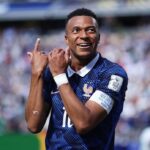 Celebrating footballer Mbappe in a blue kit on a crowded stadium making a hand gesture with his arms raised in celebration after scoring against Senegal  trybalnews