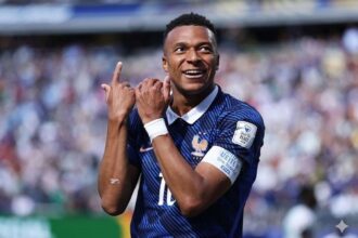 Celebrating footballer Mbappe in a blue kit on a crowded stadium, making a hand gesture with his arms raised in celebration after scoring against Senegal.