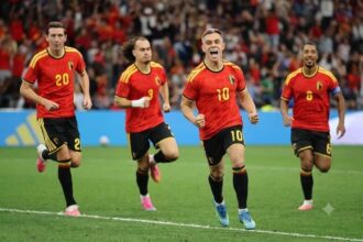 Belgium forward Leandro Trossard, wearing a red home kit with yellow shoulder stripes and number 10, celebrates with his mouth open and fists clenched after scoring. He runs on the pitch at BC Place with teammates #20, #3 with a white wrist bandage, and captain #6 with an armband behind him. All wear red shirts, black shorts with yellow numbers, and colorful cleats. 2026 FIFA World Cup Group G match between New Zealand and Belgium on June 26, 2026 at BC Place in Vancouver, British Columbia. A blurred crowd in red fills the stadium background.