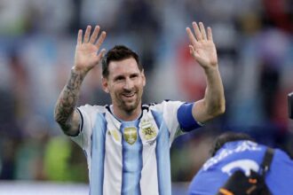 Argentina footballer in a blue-and-white striped kit raises both hands in celebration to the crowd.