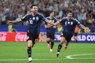 Argentina captain Lionel Messi, wearing a dark blue patterned kit with number 10 and a blue captain’s armband, celebrates with both arms outstretched and a smile. Teammates in matching dark blue kits run in behind him on the pitch. The 2026 FIFA World Cup Group J match between Jordan and Argentina took place on June 27, 2026 at Dallas Stadium in Arlington, Texas. A packed stadium crowd and red and blue advertising boards are blurred in the background.