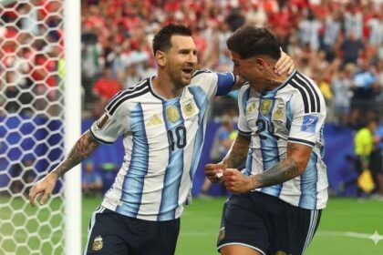 Argentina captain Lionel Messi #10 and teammate Enzo Fernandez #24 celebrate next to the goal post. Messi has his arm around Fernandez’s head and is shouting with joy. Both wear Argentina’s light blue and white striped jerseys and navy shorts. The goal net is on the left and a blurred crowd fills the stands behind them at Dallas Stadium, June 22 2026.