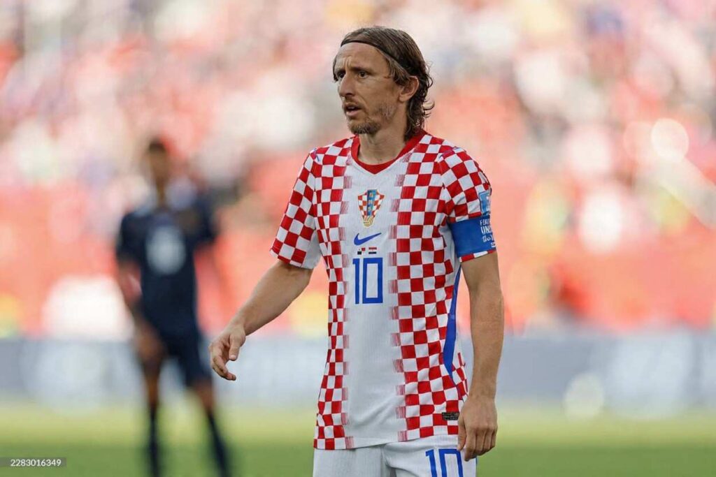 Croatia captain Luka Modrić wearing red and white checkered jersey with blue captainaposs armband stands on the pitch with a focused expression Blurred stadium crowd and opponent player in dark jersey visible in background during FIFA World Cup 2026 Group L match between Panama and Croatia at Toronto Stadium  trybalnews