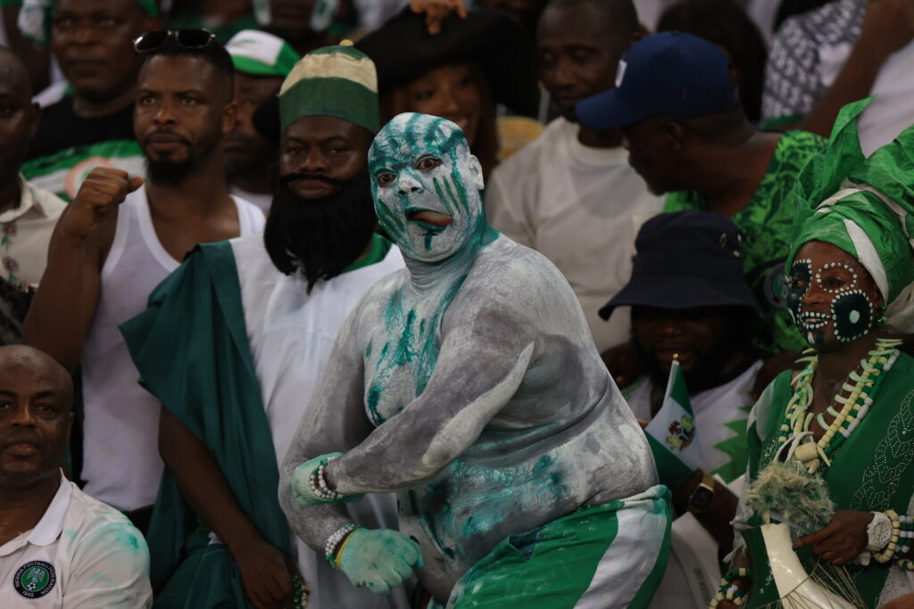 MODO1056 Trybal News Dancer in turquoise body paint performing among a crowd in green and white outfits at a stadium event with others cheering around him trybalnews