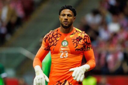 Nigerian goalkeeper in bright orange jersey with number 23, gloves on, standing on the field during a match.
