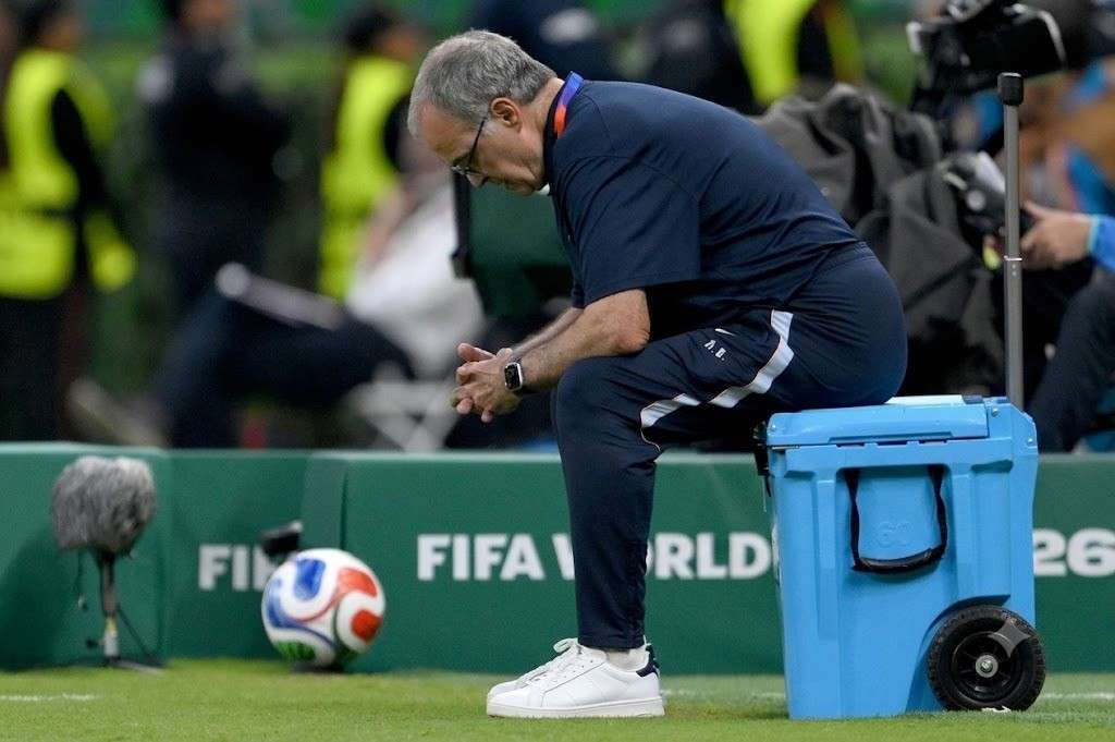 MarceloBielsa Trybal News Uruguay head coach Marcelo Bielsa a gray haired older man with glasses sits hunched forward on a blue wheeled cooler at pitchside He wears a navy Uruguay tracksuit with MB on the leg white Adidas sneakers and a black smartwatch with his hands clasped and head bowed in a dejected pose A 2026 FIFA World Cup match ball and green FIFA WORLD CUP 26 hoarding are behind him 2026 FIFA World Cup Group H match between Uruguay and Spain on June 26 2026 at Guadalajara Stadium in Zapopan Mexico Staff in yellow vests and a camera operator are blurred in the background trybalnews