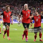 Three Austria football players in red jerseys celebrate on a green pitch as fans cheer in the stadium numbers 5 7 and 18 visible  trybalnews