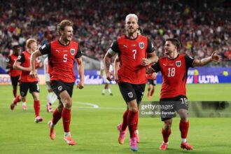Three Austria football players in red jerseys celebrate on a green pitch as fans cheer in the stadium (numbers 5, 7, and 18 visible).