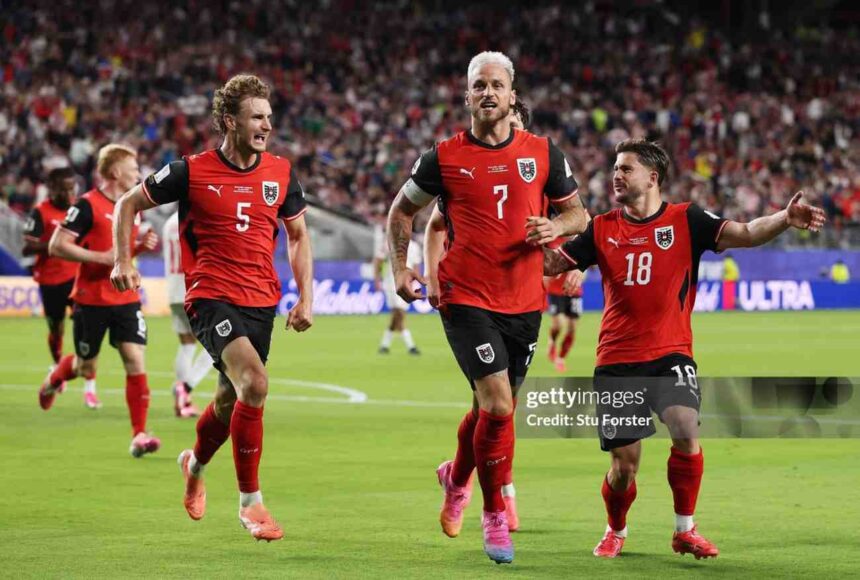 Three Austria football players in red jerseys celebrate on a green pitch as fans cheer in the stadium numbers 5 7 and 18 visible  trybalnews