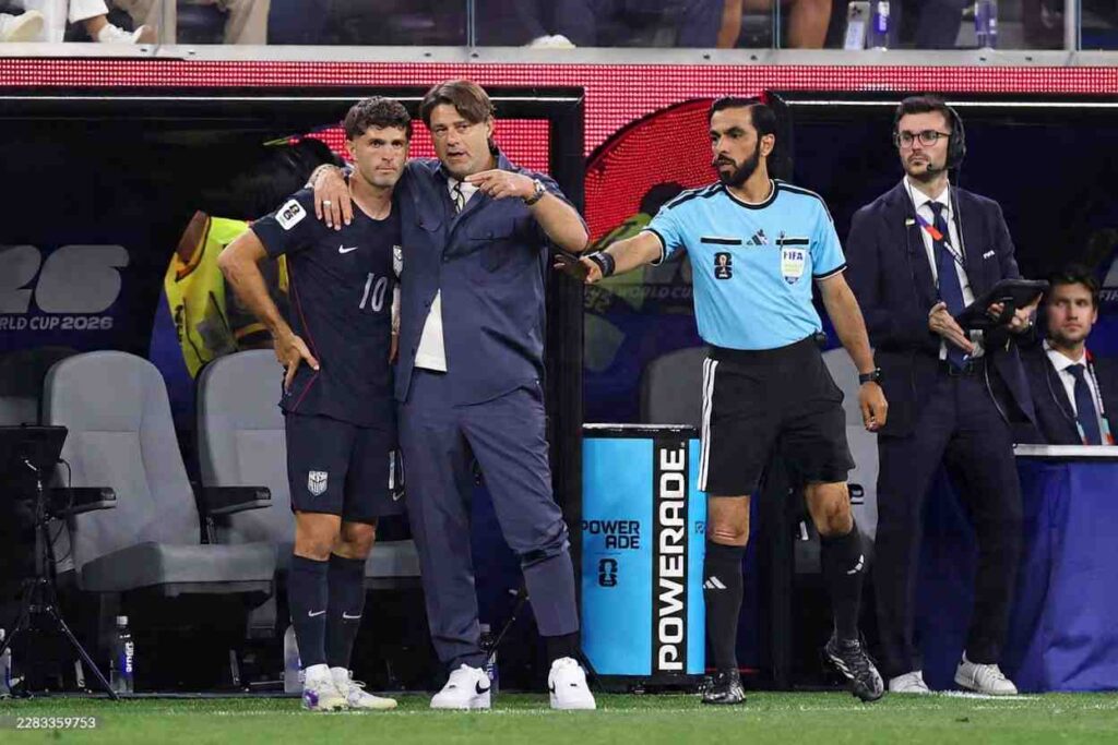 MauricioPochettinospeakstoChristianPulisic Trybal News USA Head Coach Mauricio Pochettino in a navy blue tracksuit and white sneakers stands at the touchline with his arm around midfielder Christian Pulisic 10 Pochettino points with his right hand while speaking and Pulisic in a navy USA Nike kit with 10 stands with his hand on his hip listening A fourth official in a light blue FIFA Adidas shirt and a team staff member with a headset are beside them FIFA World Cup 2026 Group D match between Türkiye and USA at Los Angeles Stadium Inglewood California on June 25 2026 USA dugout and World Cup 2026 LED boards in the background trybalnews