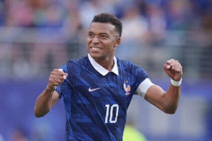 Footballer in a blue France jersey with number 10 celebrates with fists raised, smiling on the pitch.
