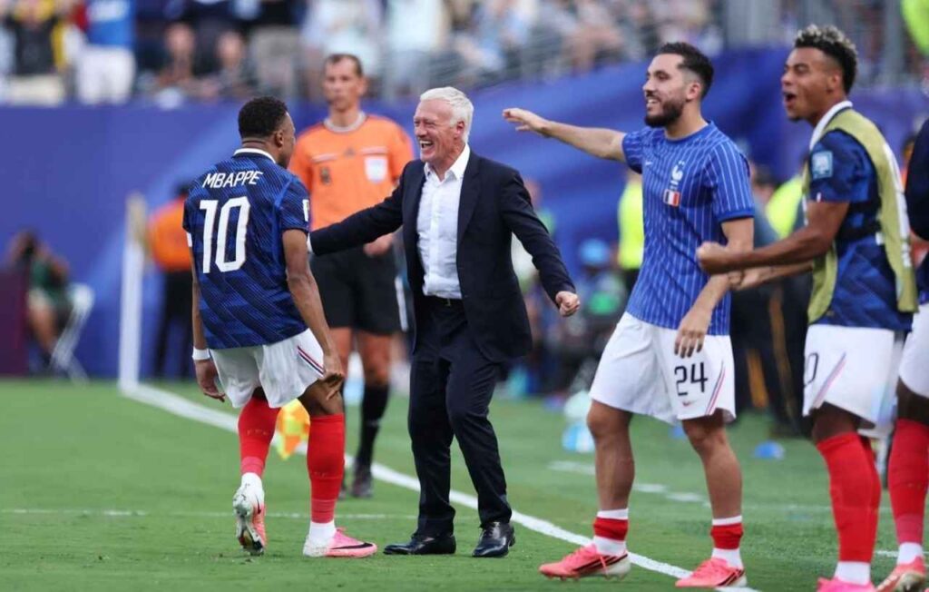 Mbappe Trybal News France forward Kylian Mbappe wearing a dark blue 10 jersey with MBAPPE on the back white shorts and red socks with pink Nike cleats celebrates with Head Coach Didier Deschamps Deschamps with white hair and a beard wears a dark suit and white shirt laughing with his arms outstretched toward Mbappe on the touchline Teammates including 24 in a blue striped jersey and another player in a green training bib celebrate nearby A referee in an orange kit stands in the background This is after Frances first goal in the FIFA World Cup 2026 Round of 32 match vs Sweden on June 30 2026 at New York New Jersey Stadium in East Rutherford New Jersey trybalnews