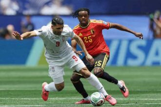 Two soccer players chase a ball during a match; white jersey number 9 versus red jersey number 25, both reaching for the ball on the field