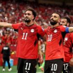 Egypt footballers Mohamed Salah wearing 10 and Mostafa Zico wearing 11 celebrate a goal Both in red Egypt jerseys with black shorts arms spread wide smiling in front of a packed red crowd at BC Place Vancouver during the FIFA World Cup 2026 Group G match vs New Zealand on June 21 2026  trybalnews