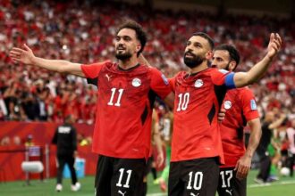 Egypt footballers Mohamed Salah wearing #10 and Mostafa Zico wearing #11 celebrate a goal. Both in red Egypt jerseys with black shorts, arms spread wide, smiling in front of a packed red crowd at BC Place Vancouver during the FIFA World Cup 2026 Group G match vs New Zealand on June 21, 2026.
