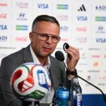 Man in a gray suit at a press conference speaking into a microphone with a soccer ball in front and sponsor logos on the backdrop behind him  trybalnews