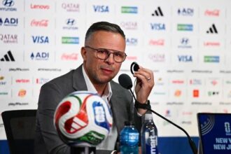 Man in a gray suit at a press conference, speaking into a microphone with a soccer ball in front and sponsor logos on the backdrop behind him.