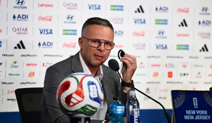 Man in a gray suit at a press conference speaking into a microphone with a soccer ball in front and sponsor logos on the backdrop behind him  trybalnews