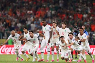 Morocco football players in white Puma kits with red numbers dash forward in celebration on the pitch. The group includes players wearing #3, #2, #26, #14, #24, #15, #16, and #9, all with arms outstretched, mouths open, and intense expressions of joy. This is after Ismael Saibari #11 scored Morocco’s fifth and winning penalty in the shootout. The FIFA World Cup 2026 Round of 32 match between Netherlands and Morocco was played on June 29, 2026 at Monterrey Stadium in Guadalupe, Mexico. A packed stadium crowd and a red Coca-Cola advertising hoarding are visible in the background.
