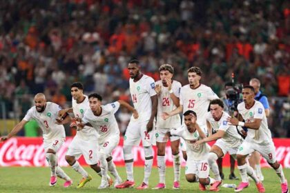 Morocco football players in white Puma kits with red numbers dash forward in celebration on the pitch. The group includes players wearing #3, #2, #26, #14, #24, #15, #16, and #9, all with arms outstretched, mouths open, and intense expressions of joy. This is after Ismael Saibari #11 scored Morocco’s fifth and winning penalty in the shootout. The FIFA World Cup 2026 Round of 32 match between Netherlands and Morocco was played on June 29, 2026 at Monterrey Stadium in Guadalupe, Mexico. A packed stadium crowd and a red Coca-Cola advertising hoarding are visible in the background.