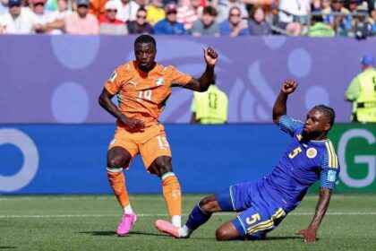 Cote d'Ivoire forward Nicolas Pepe wearing orange #19 Puma jersey, orange shorts, orange socks, and pink football boots controls the ball on the pitch. He is leaning forward with his right arm slightly out for balance as a Curacao defender in blue #5 kit slides on the ground beside him. The match ball is airborne to Pepe's left. FIFA World Cup 2026 Group E match between Curacao and Cote d'Ivoire at Philadelphia Stadium, Philadelphia, Pennsylvania on June 25, 2026. Cote d'Ivoire crest and Puma logo visible on kit.