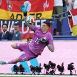 Goalkeeper in a purple kit dives to punch away a high ball during a match with flags and fans in the background  trybalnews
