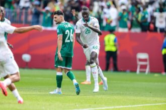 Senegal midfielder Pape Gueye #26 in a white away kit with green trim and number 26 runs forward pointing with his right index finger to celebrate. Beside him, Iraq midfielder Frans #26 in a green home kit with white number 26 walks away with his head down. Another Senegal teammate is blurred in the foreground. 2026 FIFA World Cup Group I match between Senegal and Iraq on June 26, 2026 at Toronto Stadium in Toronto, Ontario. Red stadium hoarding and fans in green Senegal shirts are visible in the background.