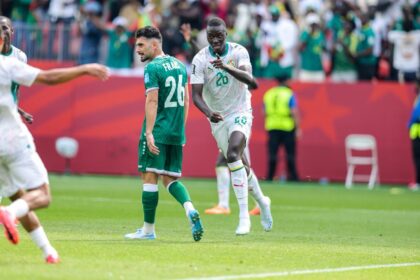 Senegal midfielder Pape Gueye #26 in a white away kit with green trim and number 26 runs forward pointing with his right index finger to celebrate. Beside him, Iraq midfielder Frans #26 in a green home kit with white number 26 walks away with his head down. Another Senegal teammate is blurred in the foreground. 2026 FIFA World Cup Group I match between Senegal and Iraq on June 26, 2026 at Toronto Stadium in Toronto, Ontario. Red stadium hoarding and fans in green Senegal shirts are visible in the background.