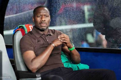 Senegal coach Pape Thiaw sits on a grey bench in the dugout wearing a brown polo shirt, black trousers, silver bracelet, and green watch. He has his hands clasped and looks intently downfield with a serious expression. Raindrops are visible on the glass in front of him. A colorful towel is draped over the seat behind him and a green jacket is next to him. The scene is before the FIFA World Cup 2026 Group I match between Norway and Senegal at New York New Jersey Stadium, East Rutherford New Jersey, June 22 2026.
