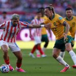 Paraguay forward Julio Enciso 19 in a red and white vertical striped Puma kit with white shorts controls the ball with his left foot Australia midfielder Jackson Irvine 22 in a gold jersey and green shorts applies pressure from the right reaching with both arms Both players have tattoos and wear bright pink boots FIFA World Cup 2026 Group D match between Paraguay and Australia at San Francisco Bay Area Stadium Santa Clara California on June 25 2026 Blurred crowd and purple advertising boards in the background  trybalnews