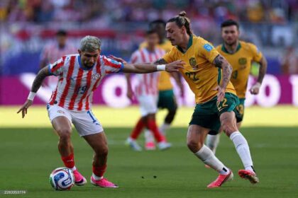 Paraguay forward Julio Enciso #19 in a red and white vertical striped Puma kit with white shorts controls the ball with his left foot. Australia midfielder Jackson Irvine #22 in a gold jersey and green shorts applies pressure from the right, reaching with both arms. Both players have tattoos and wear bright pink boots. FIFA World Cup 2026 Group D match between Paraguay and Australia at San Francisco Bay Area Stadium, Santa Clara, California on June 25, 2026. Blurred crowd and purple advertising boards in the background.