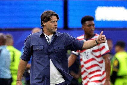 Male coach in blue outfit giving a thumbs-up on a soccer field, players and security in the background.