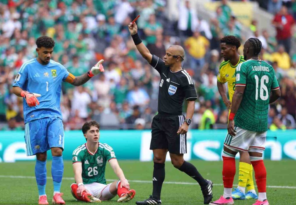 RefereeWiltonSampaio Trybal News Referee raises a red card as players stand nearby with a green team player sitting on the field and a blue clad goalkeeper watching trybalnews