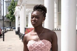 Young woman with braided hair in a pink strapless lace dress standing on a sunlit city sidewalk by white columns and a row of buildings.