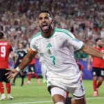 Algeria forward Riyad Mahrez wearing a white kit with green trim and number 7 celebrates with arms outstretched and mouth open after scoring Mahrez has a captains armband on his right arm and white wristbands Opposing players in red and black kits are blurred in the background on the pitch The 2026 FIFA World Cup Group J match between Algeria and Austria took place on June 27 2026 at Kansas City Stadium in Kansas City Missouri A packed stadium crowd is visible behind them  trybalnews