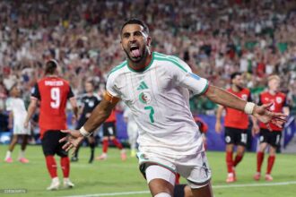 Algeria forward Riyad Mahrez, wearing a white kit with green trim and number 7, celebrates with arms outstretched and mouth open after scoring. Mahrez has a captain’s armband on his right arm and white wristbands. Opposing players in red and black kits are blurred in the background on the pitch. The 2026 FIFA World Cup Group J match between Algeria and Austria took place on June 27, 2026 at Kansas City Stadium in Kansas City, Missouri. A packed stadium crowd is visible behind them.