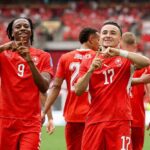 Switzerland forward Ruben Vargas wearing red 17 jersey and teammate Johan Manzambi in red 9 jersey celebrate with hand gestures forming a triangle Both players smile on the pitch with blurred red crowd and green grass in background during FIFA World Cup 2026 Group B match between Switzerland and Canada at BC Place Vancouver on June 24 2026 Switzerland defender Jaquez 2 visible behind them  trybalnews