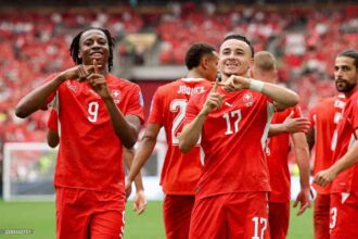 Switzerland forward Ruben Vargas wearing red #17 jersey and teammate Johan Manzambi in red #9 jersey celebrate with hand gestures forming a triangle. Both players smile on the pitch with blurred red crowd and green grass in background during FIFA World Cup 2026 Group B match between Switzerland and Canada at BC Place, Vancouver on June 24, 2026. Switzerland defender Jaquez #2 visible behind them.
