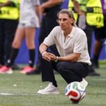 Man in a white polo crouches on a soccer field beside a ball with officials in yellow vests in the background  trybalnews