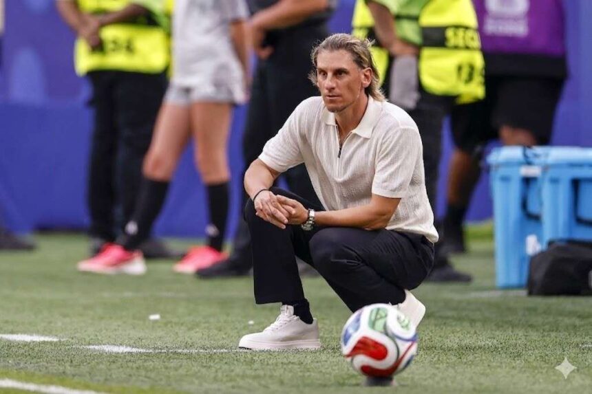 Man in a white polo crouches on a soccer field beside a ball with officials in yellow vests in the background  trybalnews
