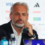 A bearded man in a suit sits at a press conference resting his chin on his hand near a microphone with sponsor logos in the blurred background  trybalnews