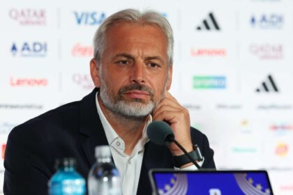 A bearded man in a suit sits at a press conference, resting his chin on his hand near a microphone with sponsor logos in the blurred background.