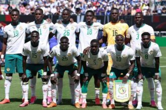 Senegal national football team posing on a sunlit pitch in white jerseys, standing and kneeling for a pre-match team photo with a banner in front.