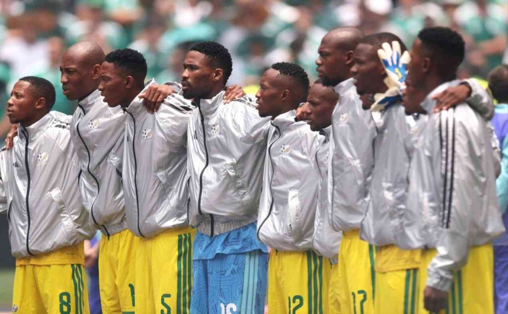SouthAfricaXI Trybal News South Africa Players stand in a row with arms around each other wearing silver jackets and yellow shorts on a field trybalnews