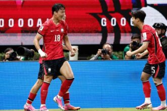 Two soccer players in red uniforms celebrate on the field as photographers film in the background.