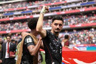 Canada midfielder Stephen Eustaquio, wearing a black speckled kit with red trim and number 7, celebrates with a raised right fist. His right wrist is wrapped in white tape. A teammate in a brown training vest puts a hand on Eustaquio’s shoulder. A large Canadian flag with a red maple leaf is visible behind them. The 2026 FIFA World Cup Round of 32 match between South Africa and Canada was played on June 28, 2026 at Los Angeles Stadium in Inglewood, California, with Canada advancing to the Round of 16. A packed crowd fills the red and blue stadium stands in the background.