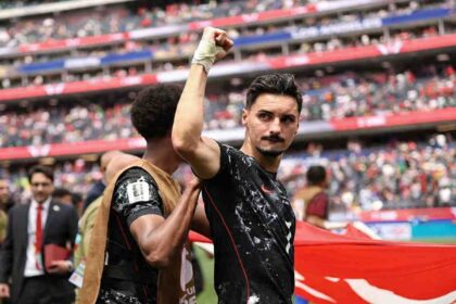 Canada midfielder Stephen Eustaquio, wearing a black speckled kit with red trim and number 7, celebrates with a raised right fist. His right wrist is wrapped in white tape. A teammate in a brown training vest puts a hand on Eustaquio’s shoulder. A large Canadian flag with a red maple leaf is visible behind them. The 2026 FIFA World Cup Round of 32 match between South Africa and Canada was played on June 28, 2026 at Los Angeles Stadium in Inglewood, California, with Canada advancing to the Round of 16. A packed crowd fills the red and blue stadium stands in the background.