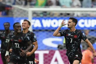 Canada midfielder Stephen Eustaquio, wearing a black kit with white speckled pattern, red trim and number 7, celebrates on the right side of frame. He raises his right hand with fingers pointing to his temple while shouting. Teammates in matching black Canada kits, including number 22, run toward him celebrating. The 2026 FIFA World Cup Round of 32 match between South Africa and Canada took place on June 28, 2026 at Los Angeles Stadium in Inglewood. A "FIFA WORLD CUP" advertising board and goal net are visible in the background.