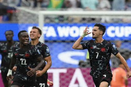 Canada midfielder Stephen Eustaquio, wearing a black kit with white speckled pattern, red trim and number 7, celebrates on the right side of frame. He raises his right hand with fingers pointing to his temple while shouting. Teammates in matching black Canada kits, including number 22, run toward him celebrating. The 2026 FIFA World Cup Round of 32 match between South Africa and Canada took place on June 28, 2026 at Los Angeles Stadium in Inglewood. A "FIFA WORLD CUP" advertising board and goal net are visible in the background.