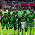 Nigeria national football team players pose for a group photo on the pitch green kits with the goalkeeper in orange on the left crowd in the stands behind them  trybalnews